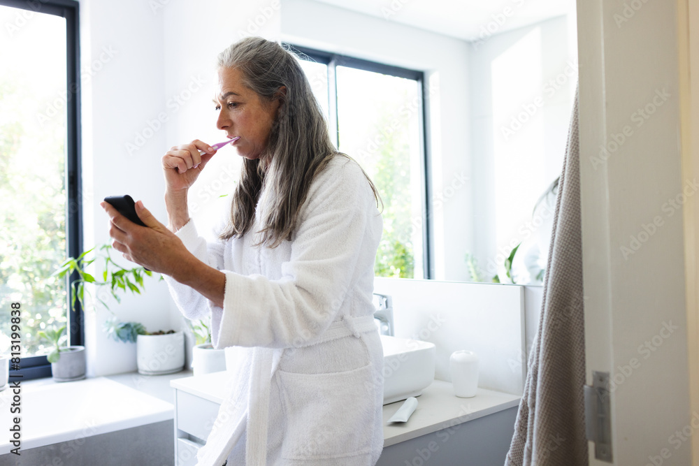 At home, mature Caucasian woman brushing teeth, checking smartphone At home, mature Caucasian woman brushing teeth, checking smartphone