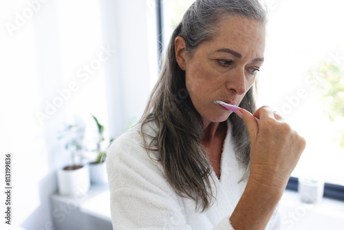 At home, mature Caucasian woman brushing teeth in a white robe