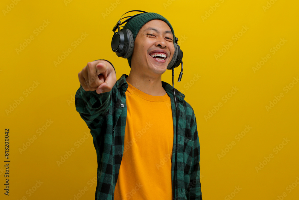 Excited Asian man in a beanie and casual clothes pumps his fist towards the camera, energized by the music playing on his headphones. Isolated on a yellow background.