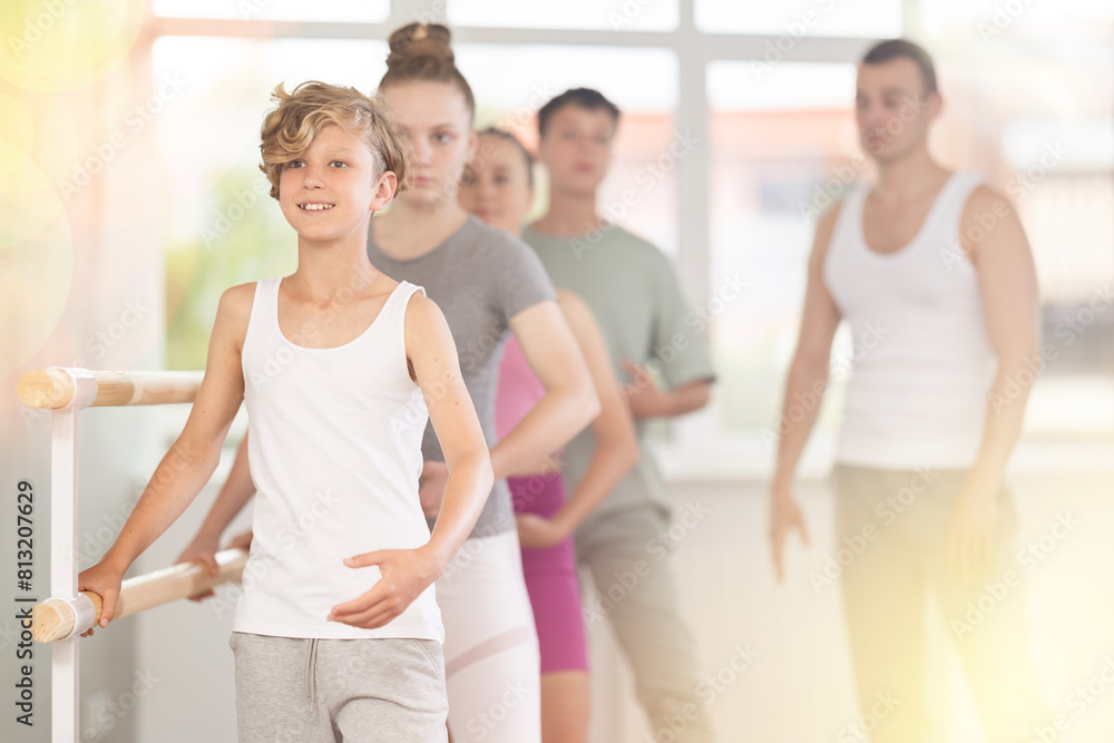 Fototapeta premium Teen boy participate in training, performs exercises, third position during group lesson. Young students engaged in ballet studio, perform repetitions of elements under male teacher guidance.