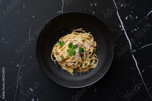 Cenital shot of a spaguetti pasta carbonara in a sleek, black ceramic bowl, set against a black marble table background, with some spices and garlic. 
