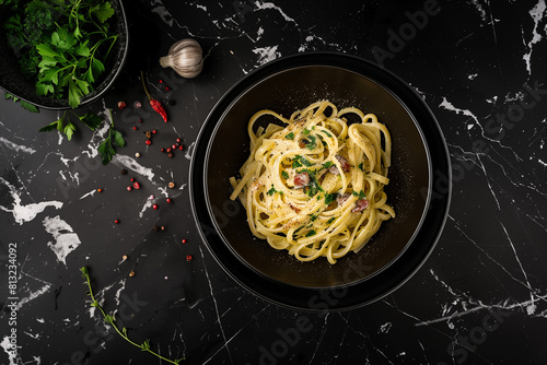 Cenital shot of a spaguetti pasta carbonara in a sleek, black ceramic bowl, set against a black marble table background, with some spices and garlic. 