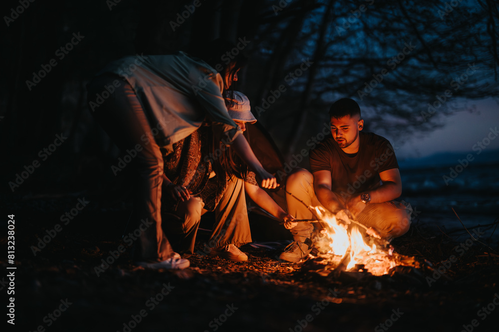 Group of friends preparing food over a campfire by the lake, embracing togetherness in nature at dusk.