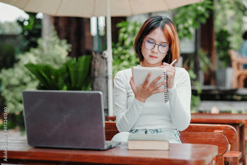 Woman in glasses is engrossed in writing notes in her notebook next to her laptop at an outdoor table. Woman in glasses is engrossed in writing notes in her notebook next to her laptop at an outdoor table.