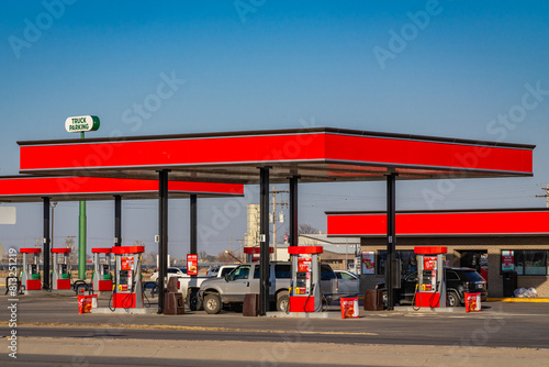 Gas station with bright red overhead roof on sunny day and cars and pick up trucks filling up with gas.