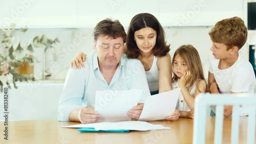 Thoughtful father considering the bills sitting at the table in the kitchen, his family members standing behind and looking at him with empathy  photo