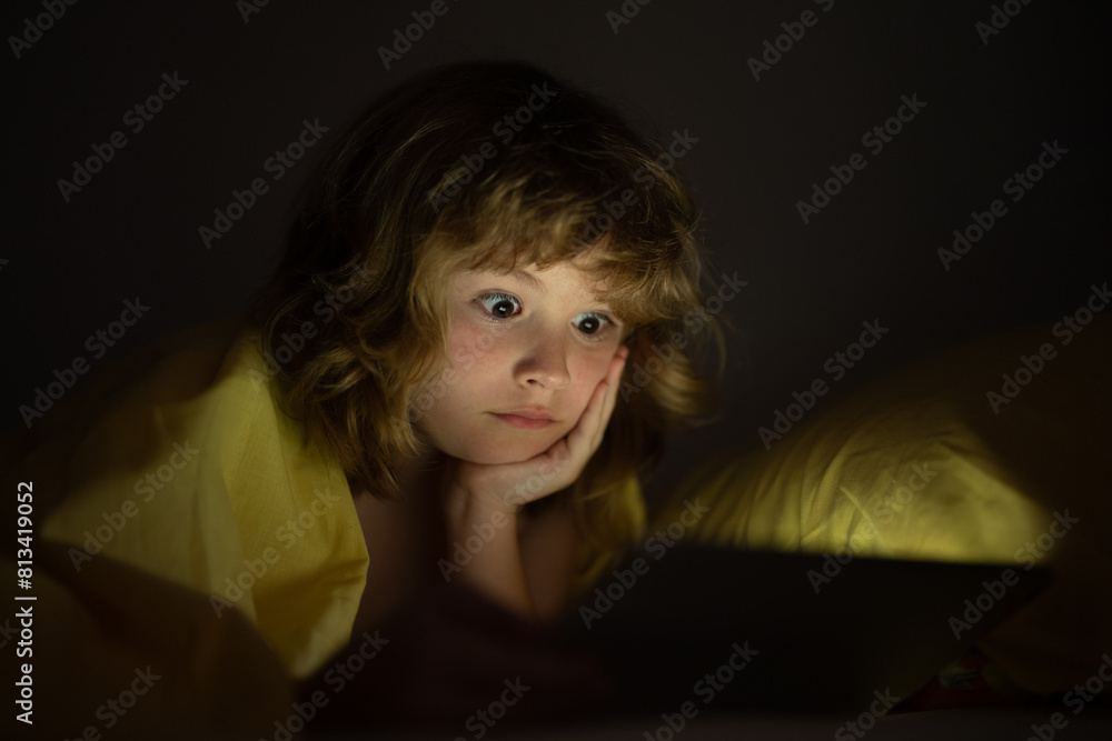 Kid is addicted to tablet, kid use tablet, watching cartoon. Child watching his tablet in the bed. Illuminated child face from device screen. Boy under the covers hold a tablet. Night time. Kid is addicted to tablet, kid use tablet, watching cartoon. Child watching his tablet in the bed. Illuminated child face from device screen. Boy under the covers hold a tablet. Night time.