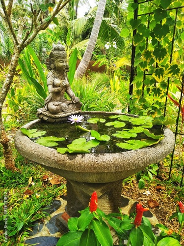 Vintage stone fountain with kneeling figure and floating lotus plants in a tropical garden with red flowers in front