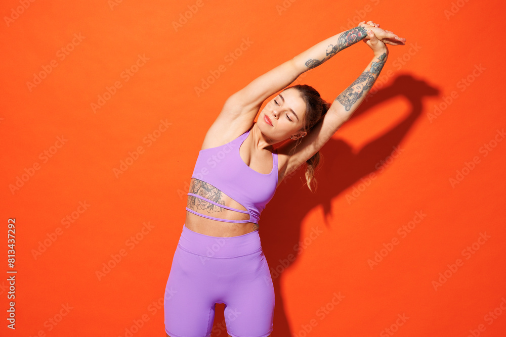Young fitness trainer sporty woman sportsman wear purple top clothes spend time in home gym do stretch exercise tilt body look aside isolated on plain orange background Workout sport fit abs concept. Young fitness trainer sporty woman sportsman wear purple top clothes spend time in home gym do stretch exercise tilt body look aside isolated on plain orange background Workout sport fit abs concept.