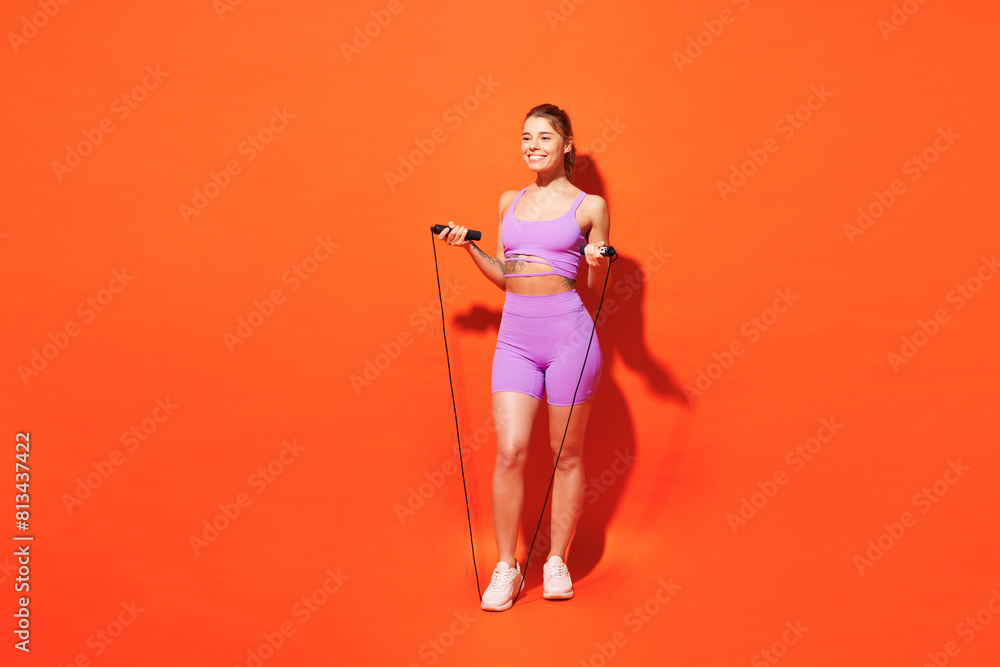 Full body happy young fitness trainer sporty woman sportsman wearing purple top clothes in home gym hold use skipping rope look aside isolated on plain orange background Workout sport fit abs concept Full body happy young fitness trainer sporty woman sportsman wearing purple top clothes in home gym hold use skipping rope look aside isolated on plain orange background Workout sport fit abs concept