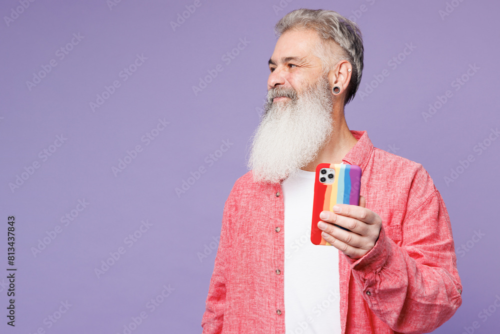 Young elderly bearded gay man 50s year old wears pink shirt casual clothes hold use mobile cell phone in striped case look aside isolated on plain purple background. Lifestyle LGBT June pride concept Young elderly bearded gay man 50s year old wears pink shirt casual clothes hold use mobile cell phone in striped case look aside isolated on plain purple background. Lifestyle LGBT June pride concept