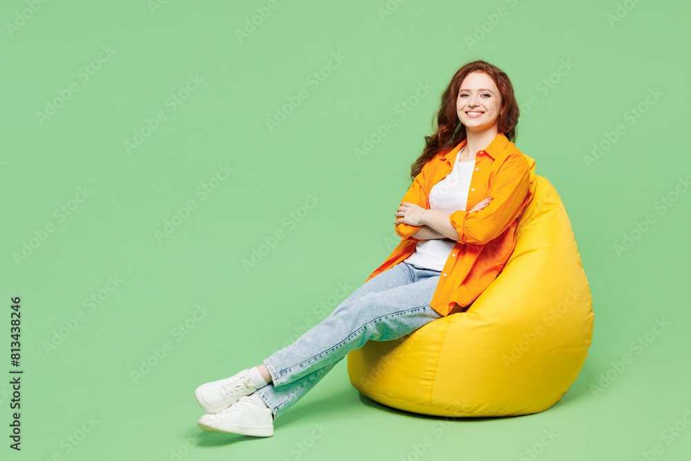 © ViDi Studio - Full body happy cheery pretty young ginger woman wear orange shirt white t-shirt casual clothes sit in bag chair rest look camera isolated on plain pastel light green background. Lifestyle concept