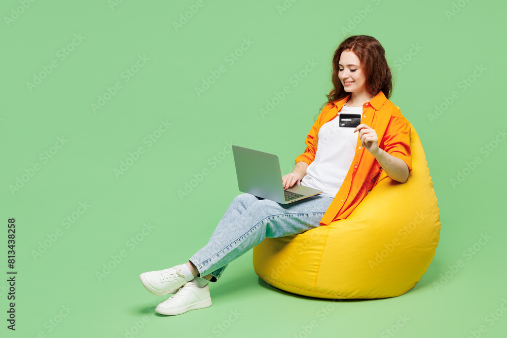 © ViDi Studio - Full body young IT woman wears orange shirt t-shirt casual clothes sit in bag chair using laptop pc computer hold credit bank card shopping online isolated on plain green background Lifestyle concept
