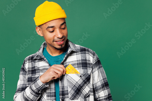 Young man of African American ethnicity he wears shirt blue t-shirt yellow hat hold put mock up of credit bank card into pocket isolated on plain green background studio portrait. Lifestyle concept.