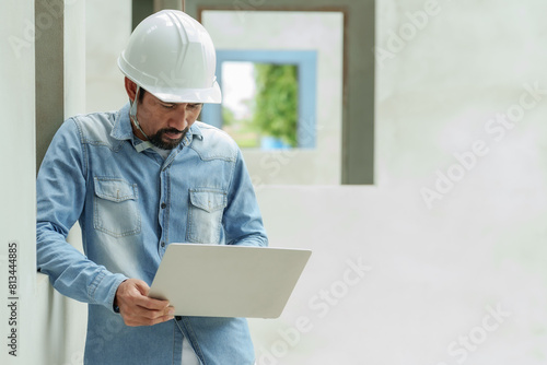 Man in blue denim shirt and white safety helmet studies laptop screen, concentration visible, standing inside construction area. Work of construction engineers on the job site