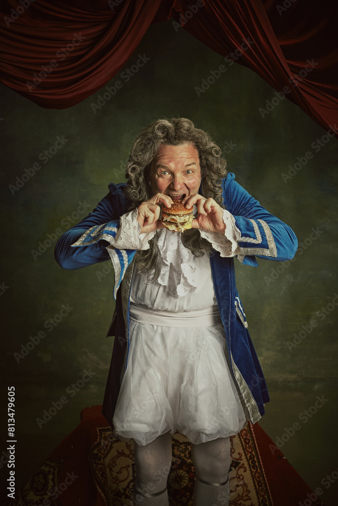 Fototapeta premium Hungry elderly man, dressed as king in classical baroque style clothes eating modern hamburger against vintage background. Concept of comparisons of eras, fusion of modernity and history, food, drink.