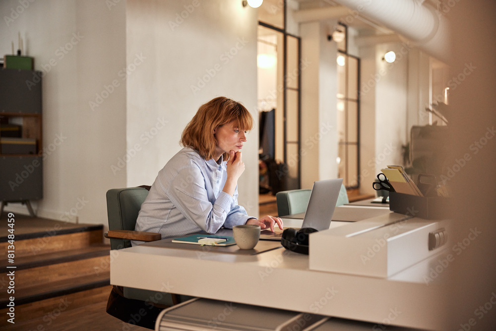 A business woman with red hair and casual work clothes, sitting focused and reading something on her laptop in the evening A business woman with red hair and casual work clothes, sitting focused and reading something on her laptop in the evening