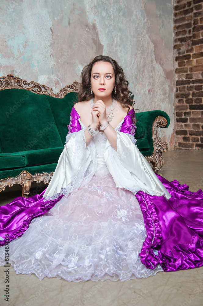 Fototapeta premium Sad beautiful woman in fantasy white and purple rococo style medieval dress sitting on the floor near sofa and praying