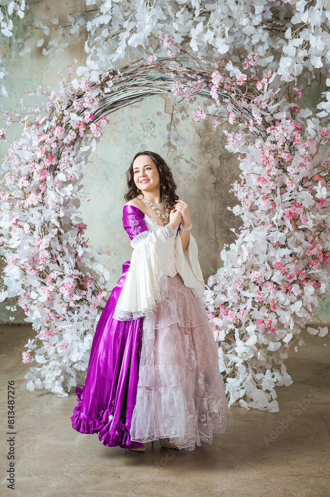 Fototapeta premium Beautiful woman in fantasy white and purple rococo style medieval dress standing near decoration with white flowers