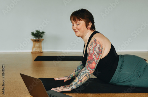 Smiling body positve young woman with tattoos streaming a yoga video on a laptop while lying on an exercise mat on the floor of a studio. She has short hair and is wearing a black tank top photo