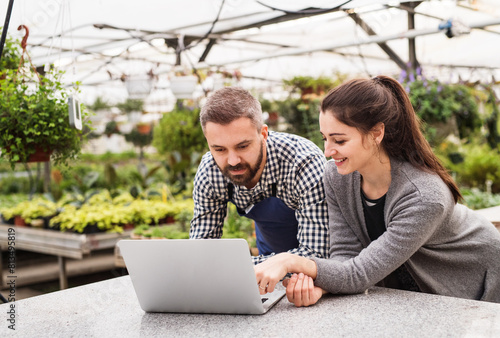 Female business owner and gardener looking at order in laptop, preparing flowers and seedlings for customer. Small greenhouse business. photo