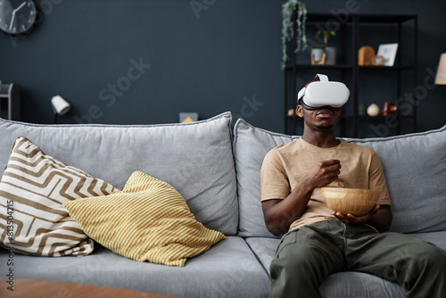 Young African American man wearing VR headset sitting on sofa in living room at home eating snacks an watching movie, copy space