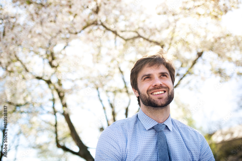Handsome businessman sitting on bench in city park. blooming tree above him, waiting for business meeting. Manager smiling, outdoor in urban setting Handsome businessman sitting on bench in city park. blooming tree above him, waiting for business meeting. Manager smiling, outdoor in urban setting