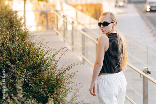 Pretty blonde girl with black sunglasses on her eyes and hair in a ponytail, wearing a black vest with a white stripe and white wide pants, standing with her back against a glass fence