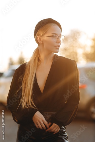Sophisticated girl with light daytime make-up and bun hair, with clear glasses on her eyes and a black beret on her head, wearing a black blouse, standing against the background of the sunset