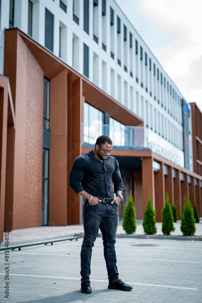 A man in a black shirt and jeans stands in front of a building. The building is a large, modern structure with a white facade.