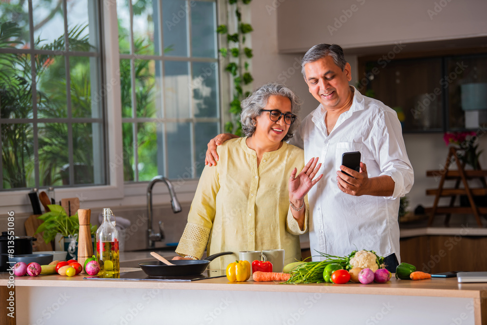 Indian asian matured retired old couple having good times cooking in kitchen and using smartphone Indian asian matured retired old couple having good times cooking in kitchen and using smartphone
