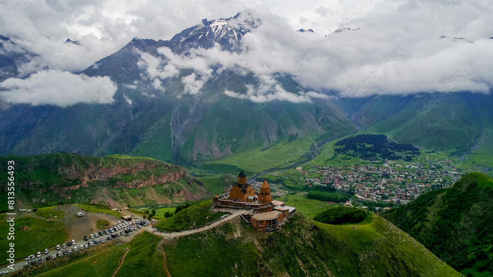 Obraz premium Aerial view of a historic monastery perched on a hilltop overlooking a valley village with snow-capped mountains and cloudy skies, possibly Georgia