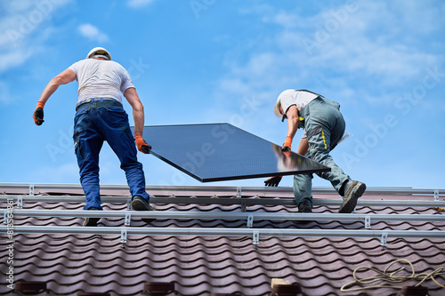 Workers building solar panel system on roof of house. Two men installers in helmets carrying photovoltaic solar module outdoors. Alternative  green and renewable energy generation concept.