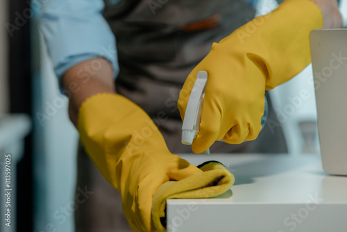 a cleaner is wiping everything in office with towel and sanitizer spray to remove all dust, wearing gloves to protect hands from chemical that use to cleanup all object for hygiene