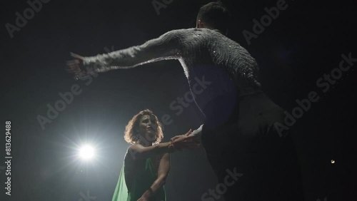 Male dancer with powerful torso in sparkling shirt rotates his girl with force and freezes in pose of toreador back to camera, backlight illumines hair and dress of girl in dark space of stage photo