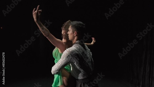 Ballroom couple dancing in darkness. Dancer in green chiton embraces her partner by neck standing with back to spectator. Man turns her by waist and pair performs support, girl on male shoulder sags photo