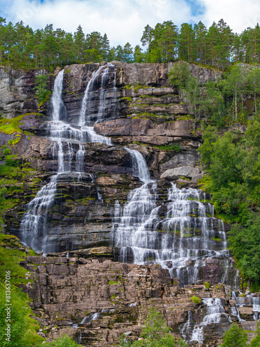 Impressive waterfall from the front. The Tvindefossen, Skulestadmo, Norway. Waterfall with several plateaus where the water falls below. The trees stand upright over the waterfall. The sky is cloudy.