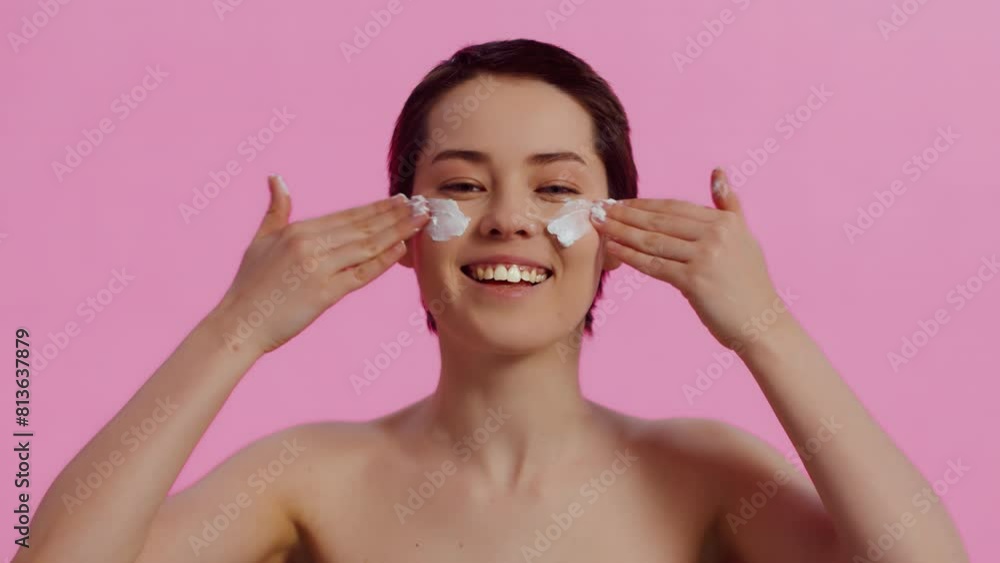 Portrait of a young beautiful woman with clean skin applying nourishing cream on her face with both hands and laughing on a pink background. Concept of skin care for health and beauty Portrait of a young beautiful woman with clean skin applying nourishing cream on her face with both hands and laughing on a pink background. Concept of skin care for health and beauty