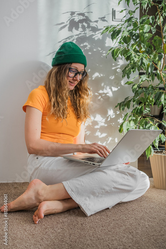 Smiling young woman with curly hair, wearing glasses and a green beanie, comfortably uses her laptop in a sunlit room surrounded by green plants.