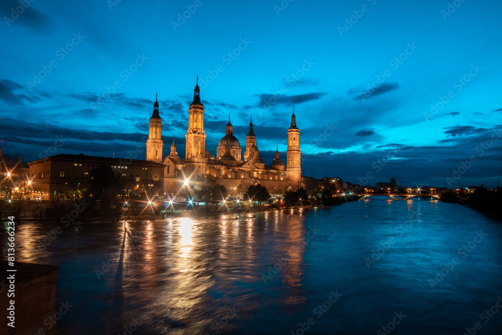 Fototapeta premium Twilight Majesty: Marveling at Zaragoza's Dusk, where the Iconic Basilica del Pilar, Stone Bridge, and Ebro River Unite in Spectacular Evening Splendor, Illuminated by the Fading Sunlight. 