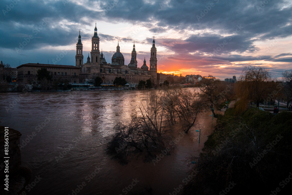 Fototapeta premium Sunset Splendor: Reveling in the Vibrant Hues of Zaragoza's Skyline as the Sun Sets in a Blaze of Orange, Casting a Spectacular Glow Across the City's Architectural Marvels and Urban Landscape. 