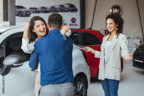 Happy couple hugging after buying a car at the dealership. Excited couple celebrating after buying a car at the dealership - car ownership concepts. © Dragana Gordic