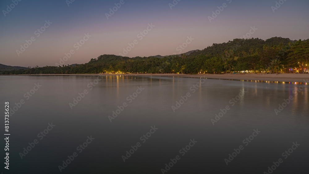 A quiet evening twilight on a tropical island. Calm ocean and sandy beach. Lights are shining among the trees on the shore. A hill against the sky, highlighted in pink. Philippines. Palawan.El nido