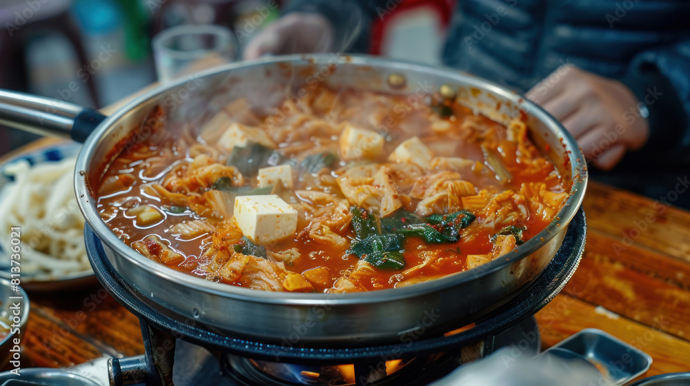 a bubbling hot pot of kimchi jjigae (kimchi stew) simmering on a portable stove atop a wooden table, filled with tender pork, tofu, and spicy fermented kimchi, emitting mouthwatering aromas. a bubbling hot pot of kimchi jjigae (kimchi stew) simmering on a portable stove atop a wooden table, filled with tender pork, tofu, and spicy fermented kimchi, emitting mouthwatering aromas.