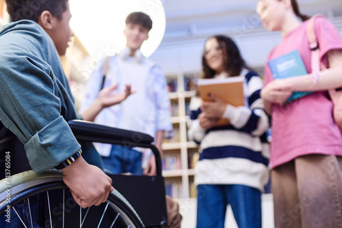 Close up of teenage boy with disability talking to group of students in school library focus on hand holding wheel copy space