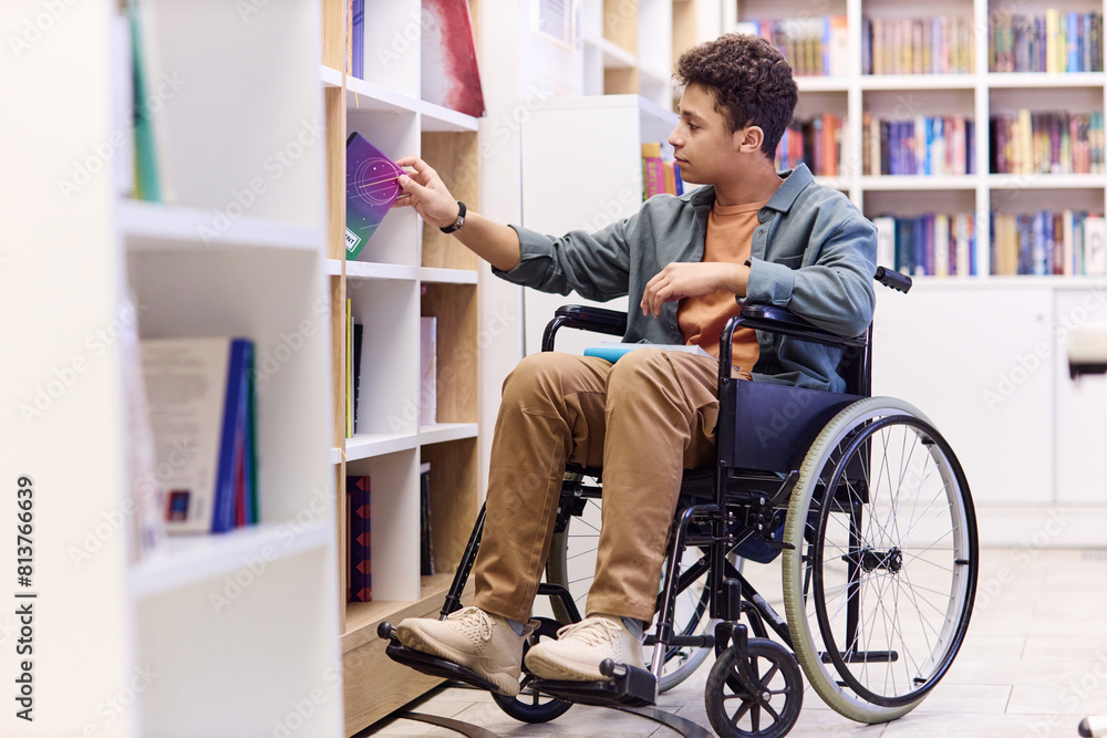 Full length portrait of Middle Eastern teenage boy using wheelchair in school library and choosing books on shelf copy space Full length portrait of Middle Eastern teenage boy using wheelchair in school library and choosing books on shelf copy space