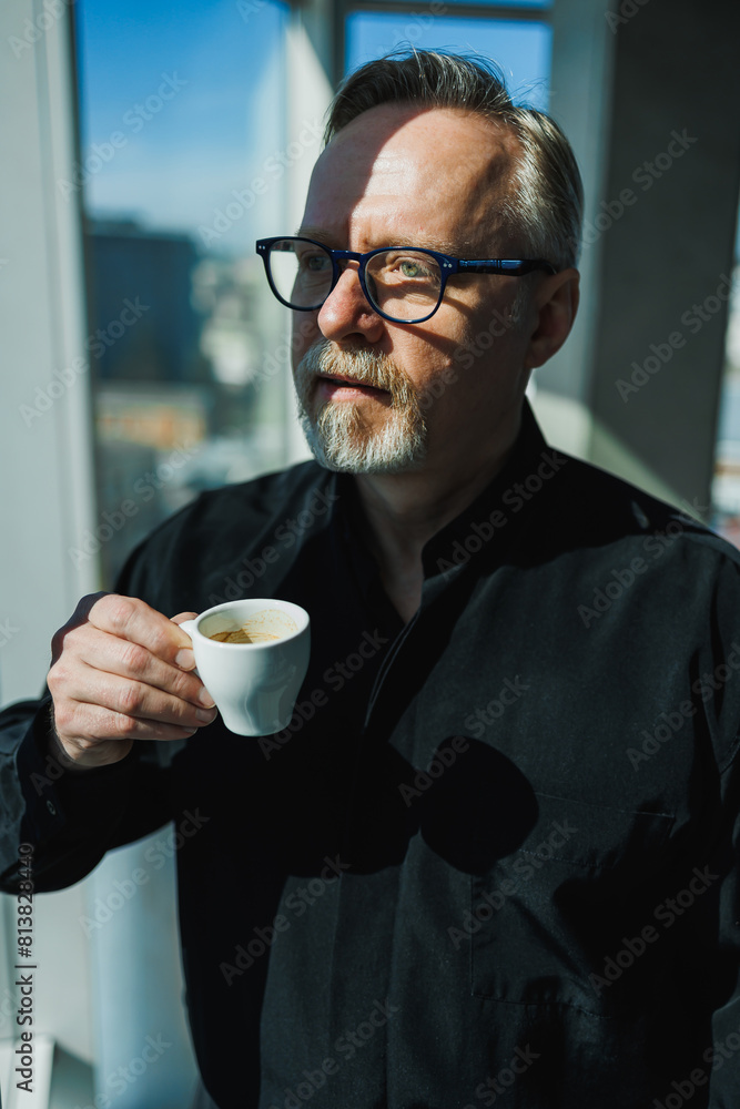 A gray-haired man in a black shirt and coffee in his hands stand by the window in the office. A gray-haired man in a black shirt and coffee in his hands stand by the window in the office.