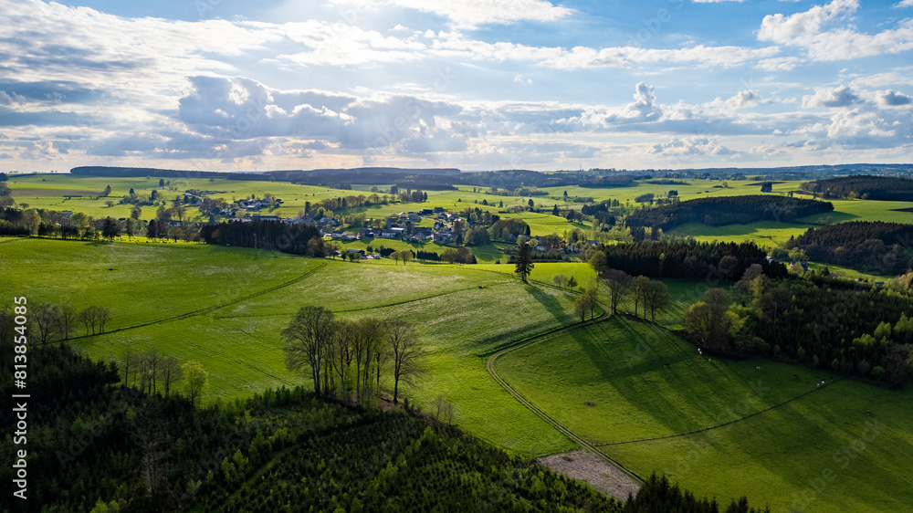 This aerial photograph captures the vast and verdant landscape of the Hautes Fagnes region, characterized by its rolling hills and vibrant green fields. The image showcases the area's natural beauty