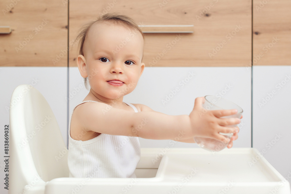 Sweet baby girl holding glass and drinking water. Happy baby sitting in a baby chair in the kitchen. Sweet baby girl holding glass and drinking water. Happy baby sitting in a baby chair in the kitchen.