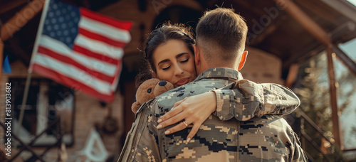 female in a military uniform hugging her husband after he came home from war, with an American flag in the background. 
